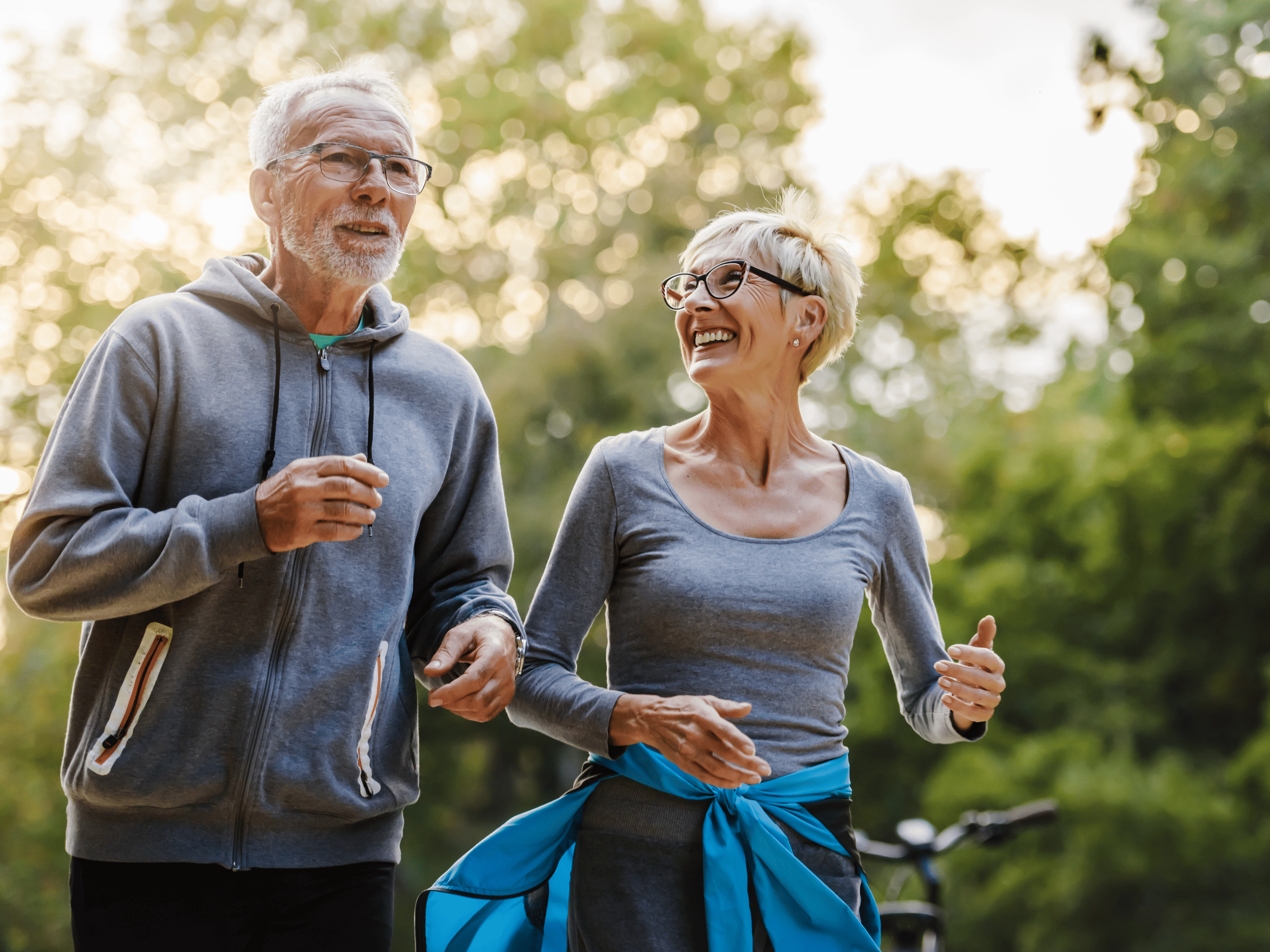 wo older adults jogging side by side on a sunny spring day, smiling and enjoying the fresh air. Lush green trees and blooming flowers line the path as they stay active and healthy together.