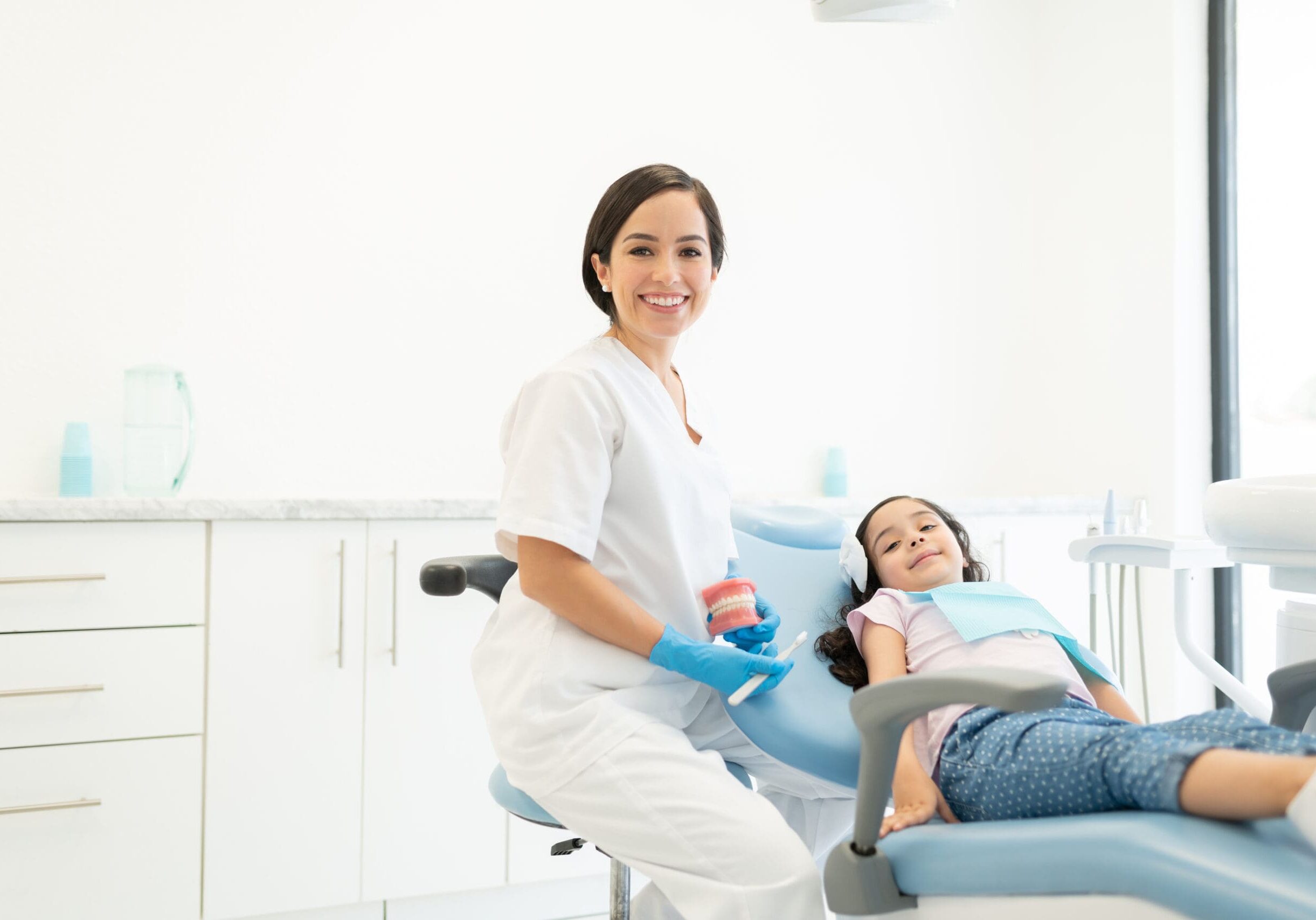 Confident female dentist with smile design model by little girl lying on chair during checkup