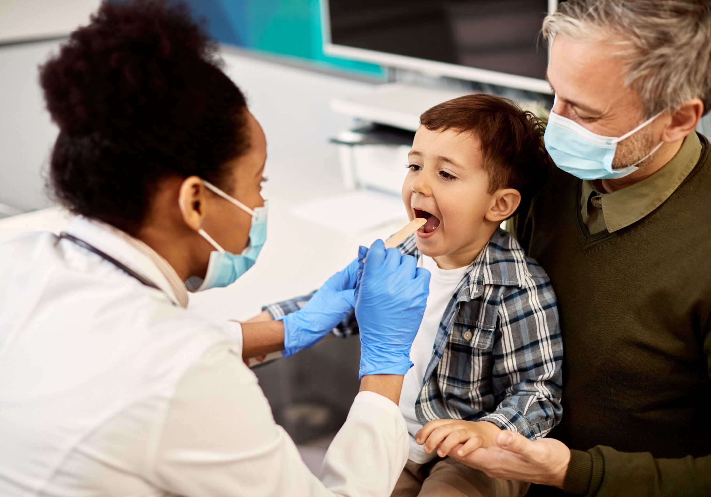 African American female dentist examining teeth of a small boy who is sitting on father's lap during dental appointment.  Focus is on boy.