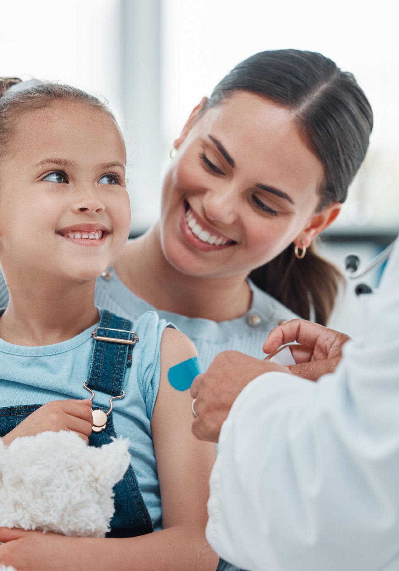 Girl, mom and doctor with vaccine injection, cotton ball and flu shot on arm for disease or covid prevention in hospital. Woman, nurse and child with pediatrician help with bandaid, teddy or health.